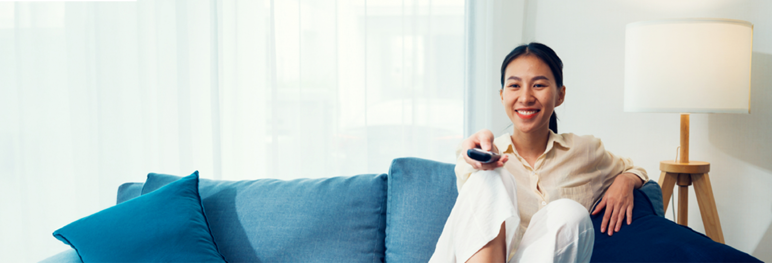 Person sitting on a blue couch in a bright living room, holding a television remote control pointed toward the viewer.