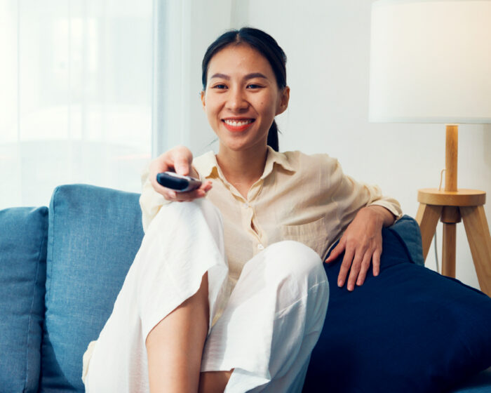 Person sitting on a blue couch in a bright living room, holding a television remote control pointed toward the viewer.